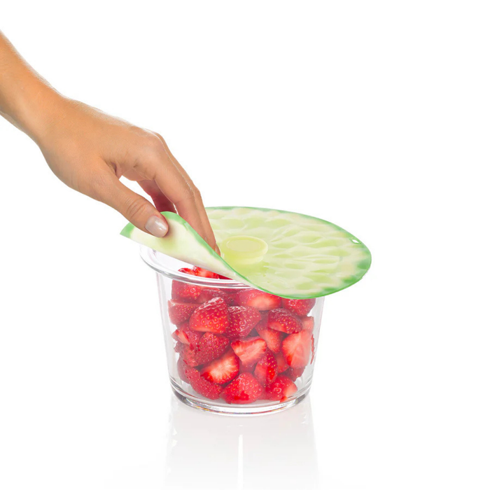 A hand is lifting a translucent silicone cup cover shaped like a cucumber slice, which is draped over a clear plastic cup filled with sliced strawberries, on a white background.