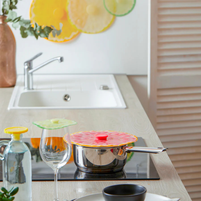 A stainless steel saucepan with a colorful citrus-patterned lid sits on a black induction cooktop in a modern kitchen setting. A wine glass and a clear bottle are in the foreground, with a sink and decorative wall art in the background.
