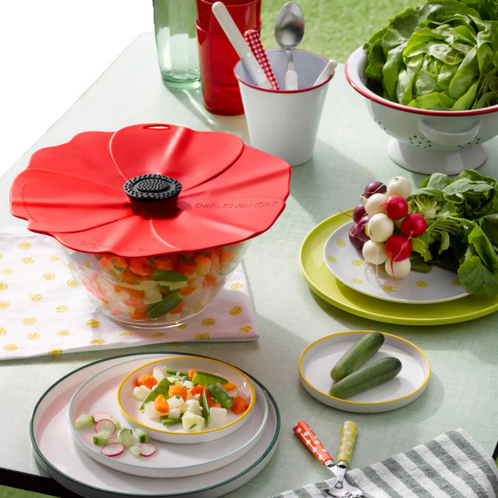 Table setting with a red poppy-shaped lid on a bowl of salad, surrounded by plates of vegetables and a salad bowl.