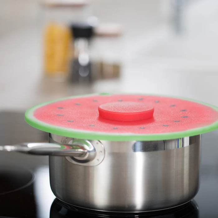 A stainless steel pot on a stovetop with a watermelon-patterned lid cover or lid itself in the foreground. The cover is bright red with a green rind-like edge and small black seed details, photographed in a blurred kitchen setting.