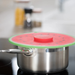 A stainless steel pot on a stovetop with a watermelon-patterned lid cover or lid itself in the foreground. The cover is bright red with a green rind-like edge and small black seed details, photographed in a blurred kitchen setting.