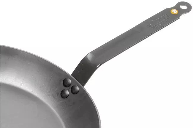 Close-up of a dark gray De Buyer Carbon Steel skillet showing the interior rim, rivets, and the long flat handle with a hanging hole and small yellow accent against a white background.