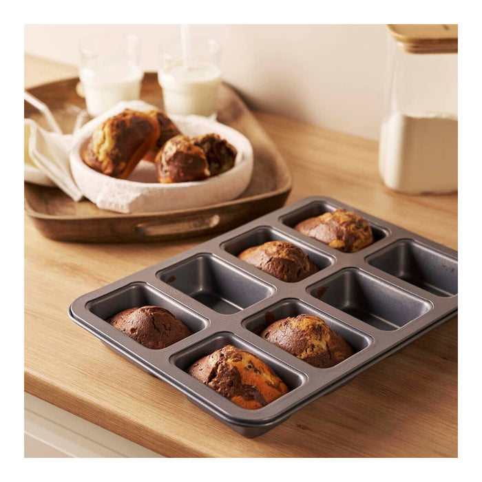 A eight-cavity mini muffin pan with baked mini muffins in the cavities, shown on a wooden countertop with a tray of more muffins and small glasses of milk in the background.