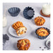 A styled food scene showing several baked muffins and a sliced muffin on a white plate, with dark fluted baking cups and milk glasses around them on a light gray surface.