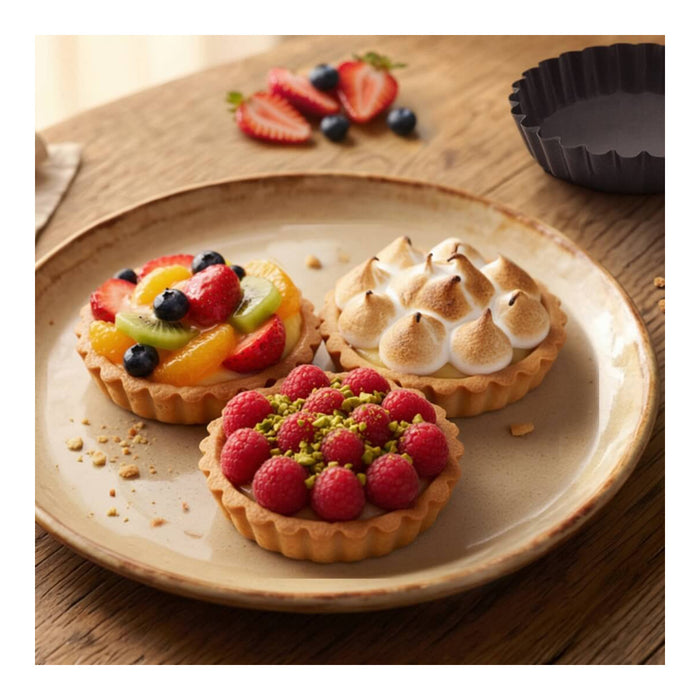 Three decorated tartlets on a beige ceramic plate, set on a wooden table. One tartlet is topped with fresh strawberries, blueberries, and kiwi slices; another has toasted meringue on top; the third is covered with fresh raspberries and chopped pistachio.