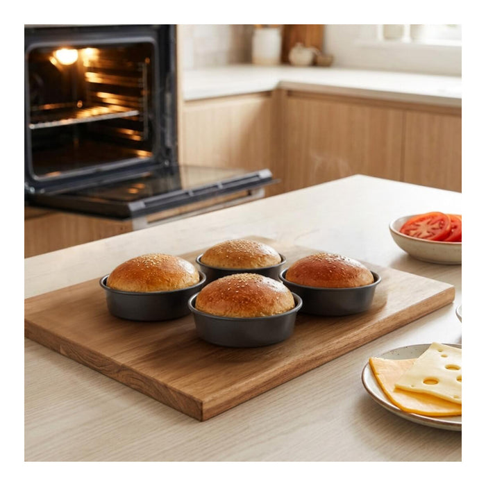 Four round dark baking pans containing golden-brown baked rolls on a wooden board, set on a kitchen counter with an oven, cheese, and tomato slices in the background.