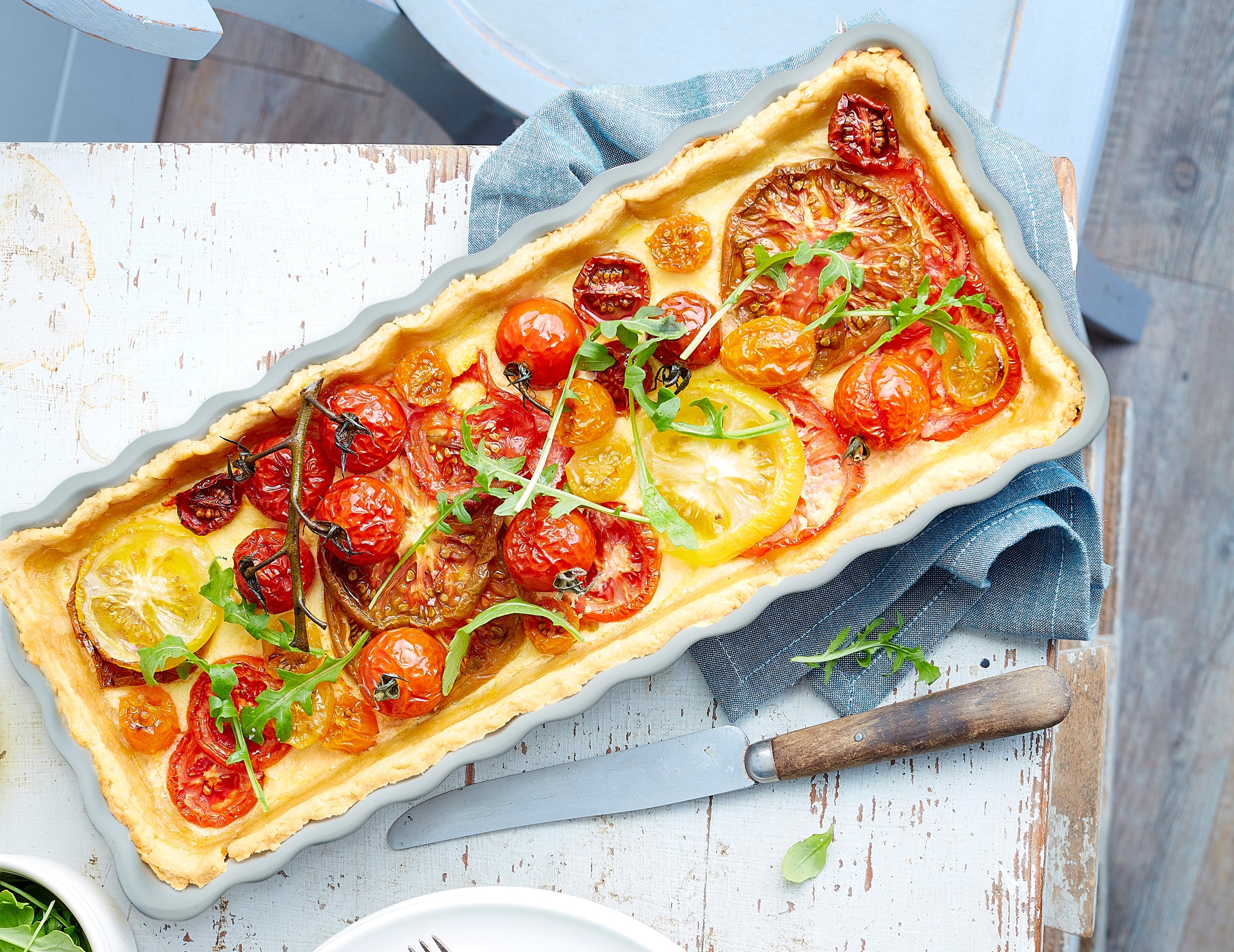 A rectangular baking dish filled with a baked tomato tart, topped with sliced yellow and red tomatoes, herbs, and a few dark olives, photographed from above on a rustic white table with a knife and napkin nearby.