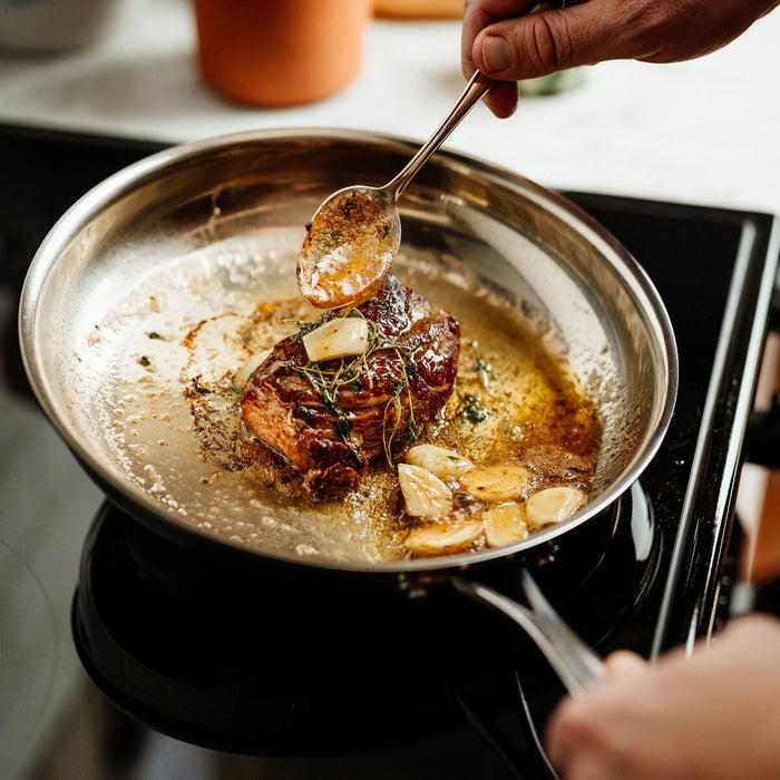 A stainless steel skillet on a stovetop with a piece of meat cooking in butter or oil, topped with garlic cloves and herbs, while a hand uses a spoon to add sauce. A blurred orange container is visible in the background.