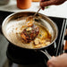 A stainless steel skillet on a stovetop with a piece of meat cooking in butter or oil, topped with garlic cloves and herbs, while a hand uses a spoon to add sauce. A blurred orange container is visible in the background.