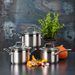 Three stainless steel pots with glass lids arranged on a dark tabletop against a textured blue-gray background, styled with a potted herb plant, a small orange pumpkin, and cooking ingredients in front.