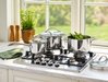 A stainless steel gas stovetop cookware set with three pots of varying sizes, glass lids, and long handles, arranged on a marble kitchen counter in front of a window.