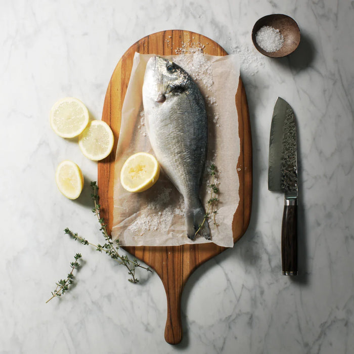 A whole raw fish on parchment paper with sea salt, arranged on a Teak serving board with lemon slices, thyme sprigs, a chef's knife, and a small bowl of coarse salt on a light marble surface.