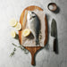 A whole raw fish on parchment paper with sea salt, arranged on a Teak serving board with lemon slices, thyme sprigs, a chef's knife, and a small bowl of coarse salt on a light marble surface.