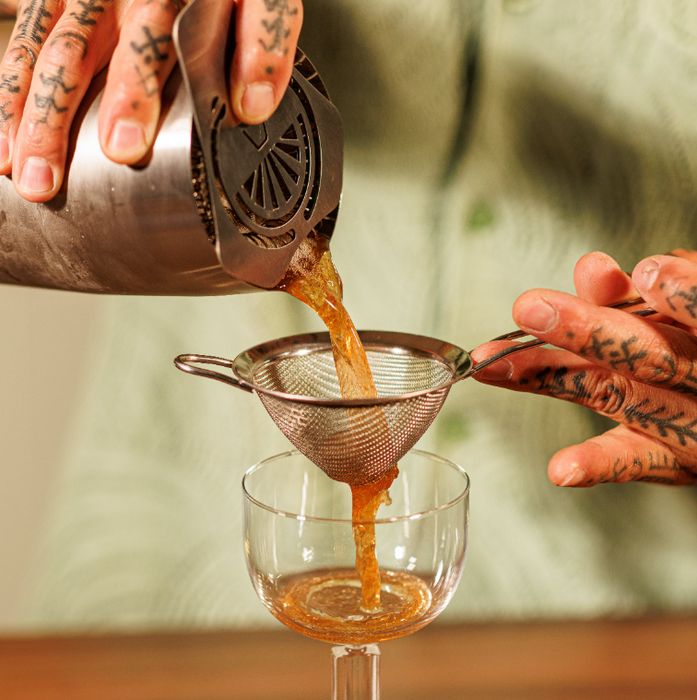 A close-up of a persons pouring an amber drink from a metal jigger, using the Vacu Vin strainer, pouring the drink into a clear coupe glass. The person's tattooed hands are visible, along with a blurred green shirt in the background.