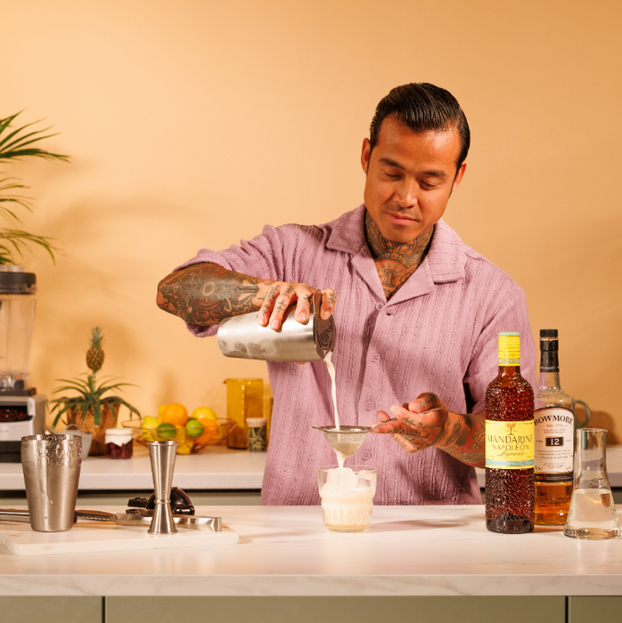 Person making a cocktail in a kitchen with bottles and ingredients on the counter, he is pouring a creamy cocktail, through the strainer into a glass that contains ice.