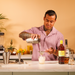 Person making a cocktail in a kitchen with bottles and ingredients on the counter, he is pouring a creamy cocktail, through the strainer into a glass that contains ice.