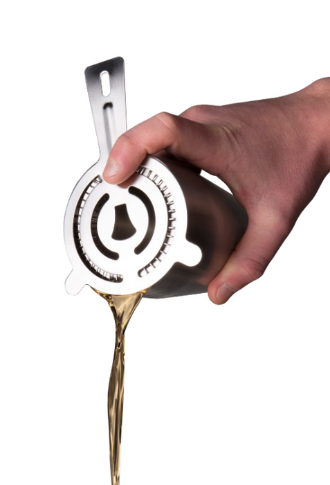 A hand holding a cocktail shaker using a Vacu Vin stainless steel cocktail strainer, pouring a golden drink, on a white background.